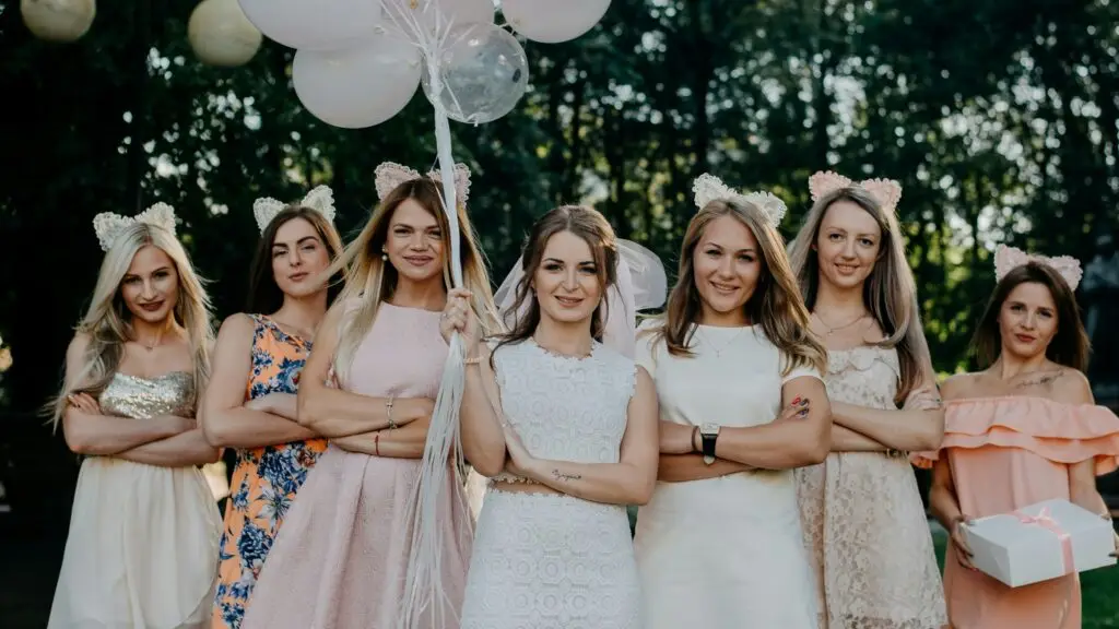 Egy menyasszony sok lufival a kezében és két oldalán egy csapat koszorúslánnyal, esküvői és elegáns ruhákban.