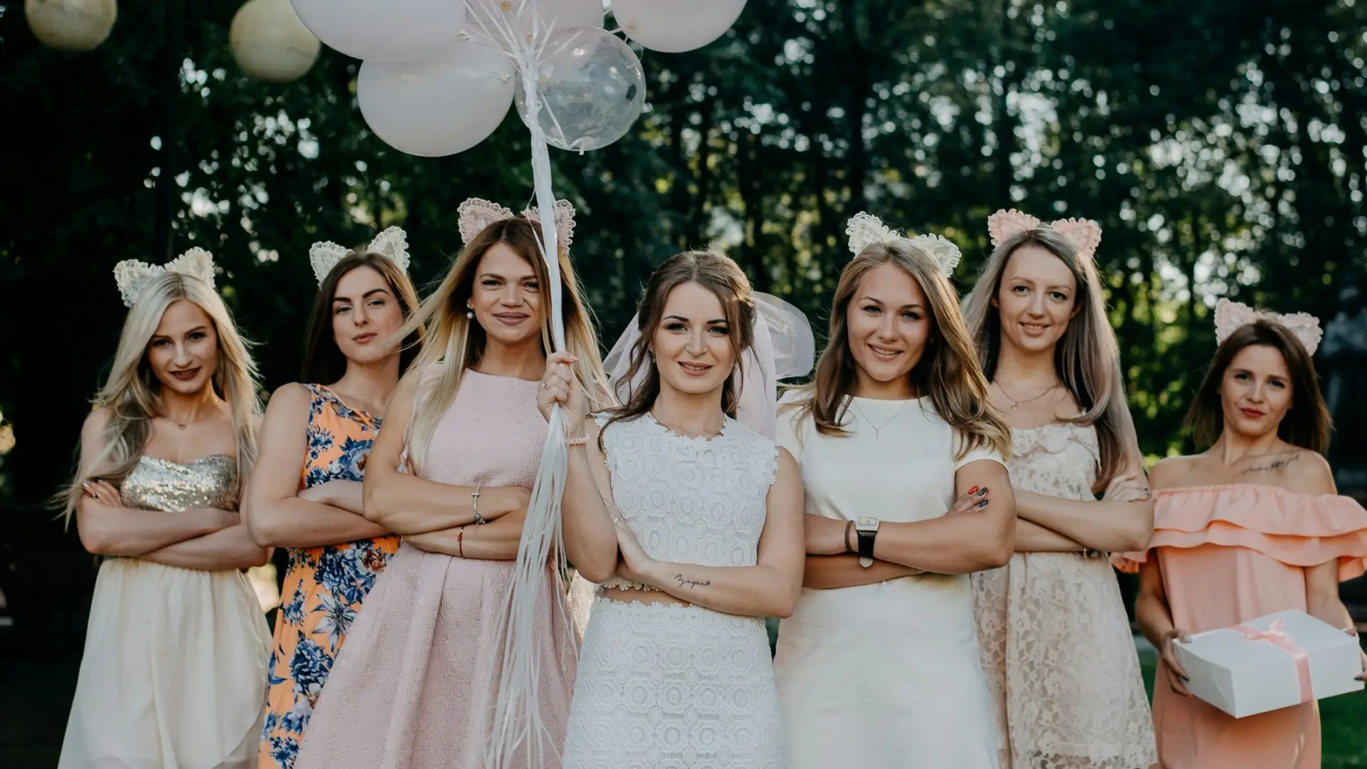 Egy menyasszony sok lufival a kezében és két oldalán egy csapat koszorúslánnyal, esküvői és elegáns ruhákban.